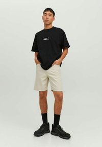 T-shirt nera con testo bianco, pantaloni corti beige, sneakers nere e calzini neri. Design semplice, vestibilità casual e tessuto leggero.