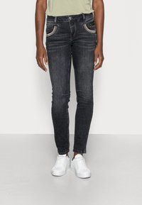 Henkilö, joka on pukeutunut mustiin slim-fit-farkkuihin, joissa on hopeiset reunukset taskuissa, ja valkoisiin lenkkareihin, seisoo tasaisella vaalealla taustalla.