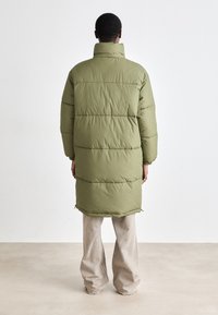 Long green puffer coat with high collar, quilted sectioning, and elastic cuffs, paired with loose beige pants. Textured fabric.