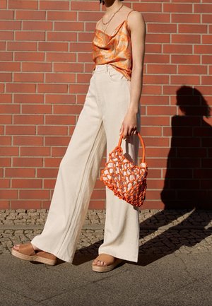 Femme portant un haut en satin à motif orange et un pantalon large couleur crème, tenant un sac à main en résille orange, debout contre un mur en briques.