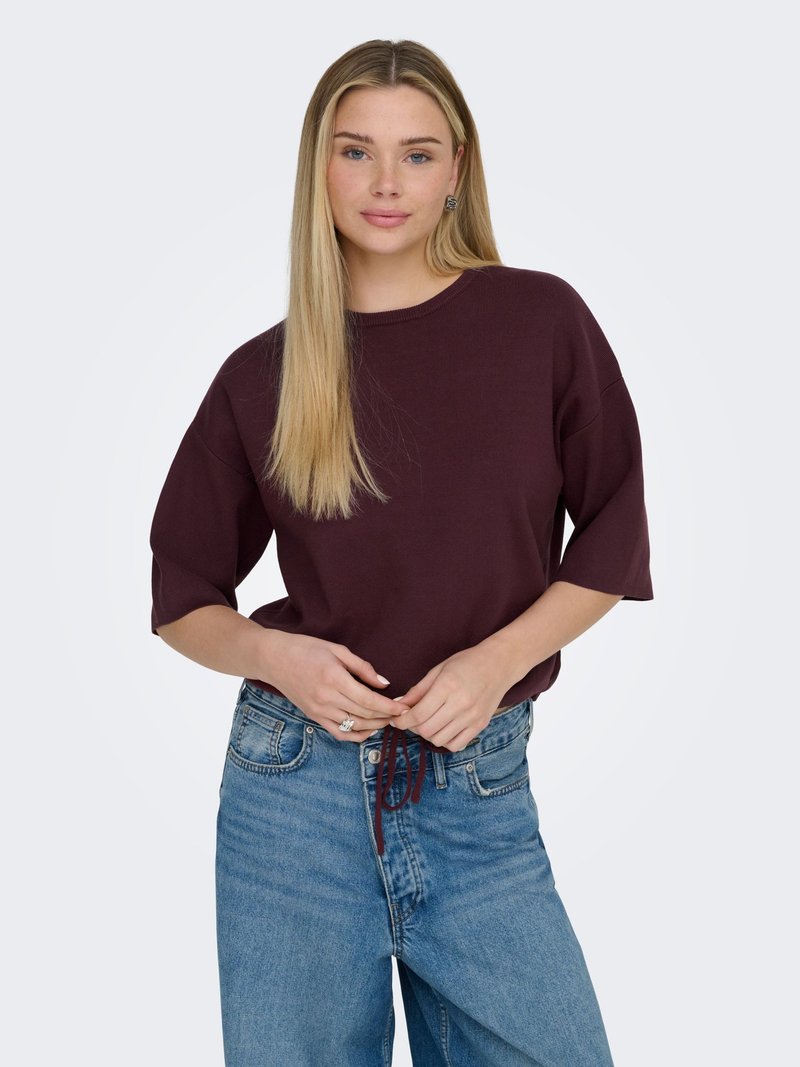 Jeune femme aux longs cheveux blonds portant un haut bordeaux à manches courtes et amples, ainsi qu'un jean bleu, tenant les cordons à la taille.