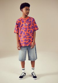 Oranje T-shirt met korte mouwen en paarse abstracte patronen, gecombineerd met knielange denim shorts en zwarte sneakers met witte accenten.