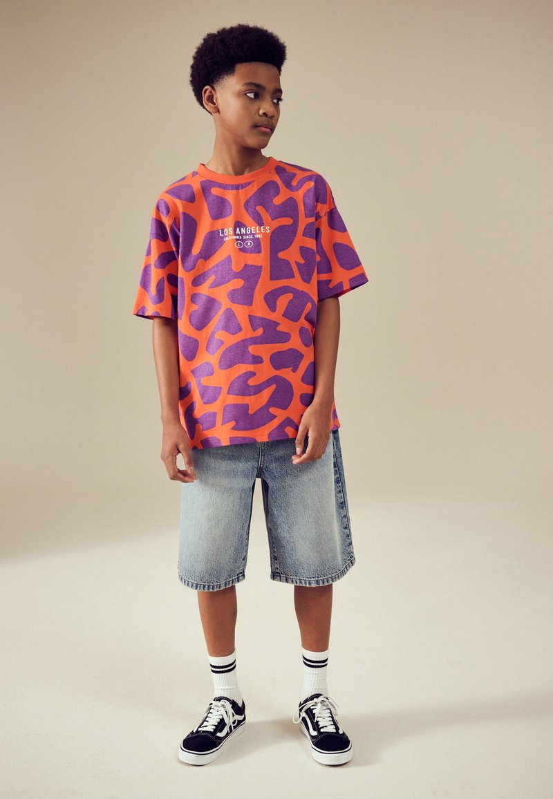 Oranje T-shirt met korte mouwen en paarse abstracte patronen, gecombineerd met knielange denim shorts en zwarte sneakers met witte accenten.
