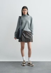 Szara bluzka z długim rękawem, brąskowa skórzana spódnica z szwami, czarna torebka na ramię, białe skarpetki oraz czarno-brązowe sneakersy z kremowymi akcentami.