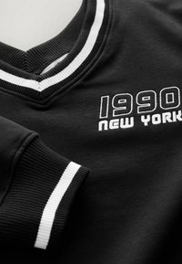 Sweat-shirt noir avec col et poignets côtelés blancs rayés, texte brodé "1990 New York" sur la poitrine.