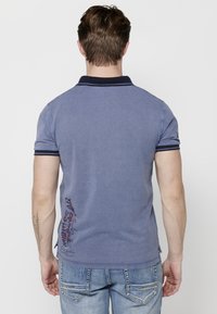 Polo majica s kratkimi rokavi v svetlo modri barvi z mornarsko modrimi detajli. Ima vezenino na hrbtni strani in stranske razpoke na robu.