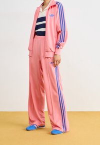 Rosa träningsoverall gjord av ett slätt tyg med blå ränder längs ärmarna och byxorna. Inkluderar en vit och marinblå randig skjorta. Ljusblå skor.