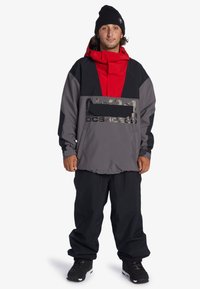 Pánska mikina s kapucňou v šedej farbe s červenými akcentmi, s prednou kapsou, kapucňou a čiernym logom. Kombinovaná s voľnými čiernymi nohavicami a obuvou.