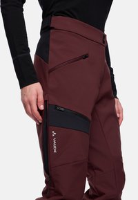 Bordeauxrode softshellbroek met zwarte accenten, voorzien van een hoge tailleband, meerdere met rits afsluitbare zakken en een gladde textuur. Vaude-logo zichtbaar.