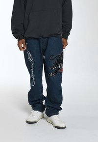 Jeans in denim con una vestibilità ampia, caratterizzati da un design ricamato di una pantera nera su una gamba e testo bianco sull'altra. Indossati con sneakers bianche.