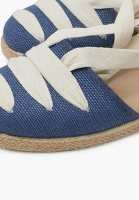 Modré tkané espadrilky s bílými textilními popruhy, s přírodní juteovou podrážkou. Design je plochý a s zaoblenou špičkou.