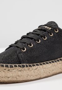 Adidași negri cu textură și strălucire metalică, având ochiuri rotunde aurii, șireturi negre și o talpă țesută din iută.