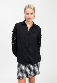 Schwarze Hemdbluse mit langen Ärmeln und strukturierten schwarzen floralen Verzierungen. Kombiniert mit einem Rock im Hahnentritt-Muster.