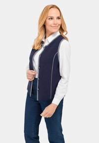 Vrouw met blond haar die een marineblauwe vest met knopen over een wit shirt en blauwe jeans draagt, waarbij ze naar de zijkant kijkt en lichtjes glimlacht.