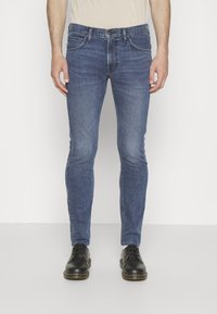 Jeans de denim azul ajustados con un acabado desgastado, que cuentan con bolsillos frontales y un cierre de botón, combinados con zapatos de cuero negros.