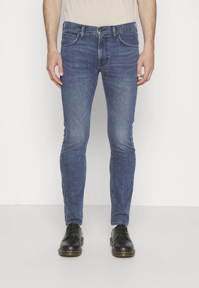 Jeans de denim azul ajustados con un acabado desgastado, que cuentan con bolsillos frontales y un cierre de botón, combinados con zapatos de cuero negros.