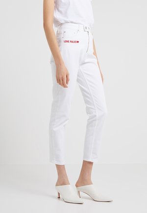 Frau trägt weiße Jeans mit hohem Bund und rotem "LOVE RULES"-Schriftzug auf der Tasche, weißen Oberteil und weiße Mules mit Absatz, stehend vor einfarbigem Hintergrund.