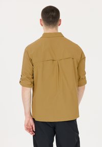 Lichtbruine shirt met korte mouwen, voorzien van een rugplooien en een plooi-detail. De mouwen zijn opgerold en de stof lijkt glad en lichtgewicht.