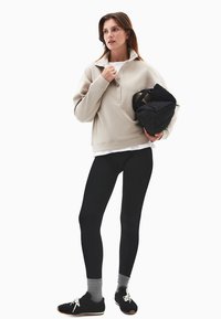 Jasno beżowy sweter z wysokim kołnierzem i detalami na guziki, czarne legginsy, szare skarpetki i czarne buty teksturowane; trzyma czarną pikowaną torbę.