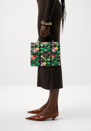 Personne portant une robe marron côtelée tient un sac à main floral avec une poignée supérieure, portant un bracelet épais en or et des chaussures marron à talons bas et pointues.