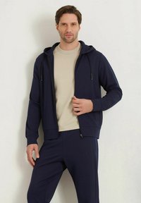 Sudadera con cremallera en color marino, con cordón ajustable, fabricada en un tejido suave y con puños acanalados; llevada sobre una camiseta de manga corta en color beis claro y pantalones a juego.