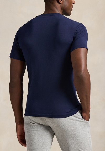 Férfi, aki testhezálló sötétkék rövid ujjú inget és világosszürke melegítőnadrágot visel, egyik kezét a zsebében tartva, hátulnézetben.