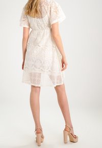 Vit spetsklänning med korta ärmar, med intrikata blommönster. En lagerdesign med genomskinlig textur, kombinerad med beige högklackade sandaler.