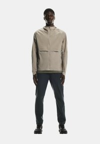 Gaisms, beigs jakas ar priekšējo rāvējslēdzēju, elastīgām piedurknēm un pieguļošu dizainu. Apvienots ar tumšiem biksēm un sportiskām kurpēm.