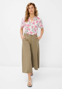 Chemise à manches courtes à motifs floraux rose, vert et bleu ; pantalon large kaki ; sandales blanches à lanières. Le modèle se tient avec les mains dans les poches.