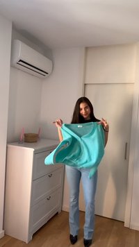 Ung kvinne holder en turkis genser i et lyst rom med hvite møbler og tregulv, og gjør seg klar til å ta den på.