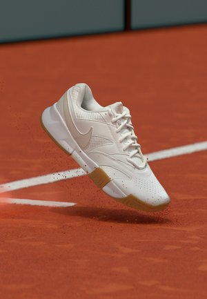 Weißer Nike-Sneaker mit Gummisohle wirbelt roten Tonstaub auf einem Tennisplatz nahe einer weißen Begrenzungslinie auf.