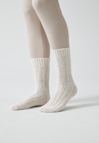 Calcetines blancos de punto con textura de canalé y un puño enrollado, llevados sobre piernas cubiertas con medias beige, sobre un fondo liso.