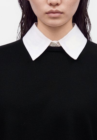Gros plan d'une personne portant une chemise blanche à col sous un pull noir, montrant le cou, le col et le bas du visage.