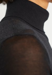 Gros plan d'une personne portant un pull à col roulé noir avec un tissu transparent sur les épaules, sur un fond neutre.