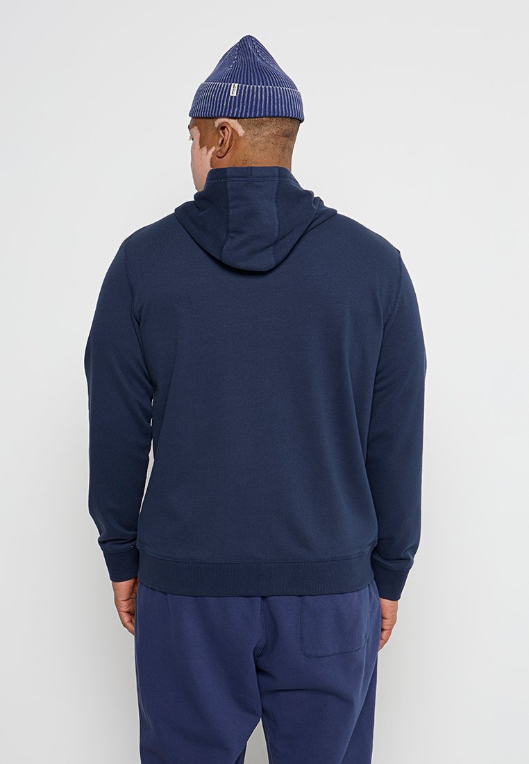 Granatowy hoodie z kapturem na sznurki, prążkowanymi mankietami i dołem. Miękka tkanina o gładkim wzorze. Noszony z dopasowanymi granatowymi spodniami.