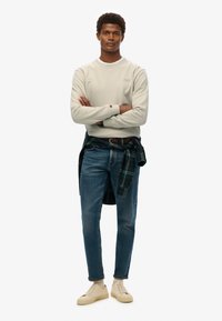 Sudadera gris claro con escote redondo, jeans azules y zapatillas beiges. Camisa de cuadros atada en la cintura, que muestra su patrón en verde oscuro y negro.