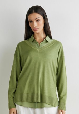 Jeune femme aux longs cheveux noirs portant un pull ample vert clair à col en V par-dessus une chemise verte à boutons, devant un mur uni.