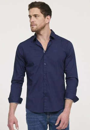 Chemise bleu marine à boutons, manches longues, coupe slim, avec un logo discret sur la poitrine et des accents blancs contrastants sur les poignets.