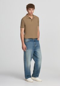 Giovane uomo in piedi con le mani nelle tasche di jeans blu sbiaditi, indossando una maglietta polo marrone e sneaker bianche su uno sfondo bianco.