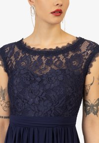 Marineblauwe jurk met een kanten bodice met bloemmotieven, een scalloped halslijn en een gladde taille. Inclusief oorbellen als accessoire.