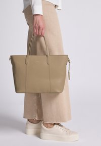 Borsa tote in pelle beige con superficie texturizzata, due manici superiori e logo dorato. Abbinata a pantaloni a gamba larga color tan chiaro e sneakers color crema.
