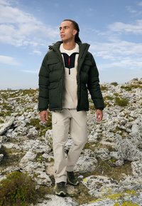 Donker groene pufferjack met rits, twee voorkruimels. Beige cargobroek, groene wandelschoenen. Buitensetting met rotsachtig terrein.