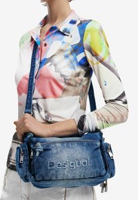 Sac bandoulière en denim bleu avec poches zippées, logo "Desigual" en relief et sangle réglable. Présente une surface texturée et des accents noués.