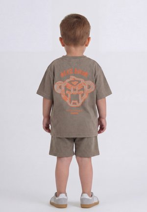 Jong kind met de rug naar de camera, draagt een grijze korte broek en een bijpassend shirt met een oranje grafisch ontwerp en tekst op de achterkant.