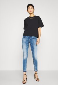 Person steht da und trägt ein lockeres schwarzes T-Shirt, hellblaue enge Jeans mit ausgefransten Saum und schwarze trägerlose High Heels.