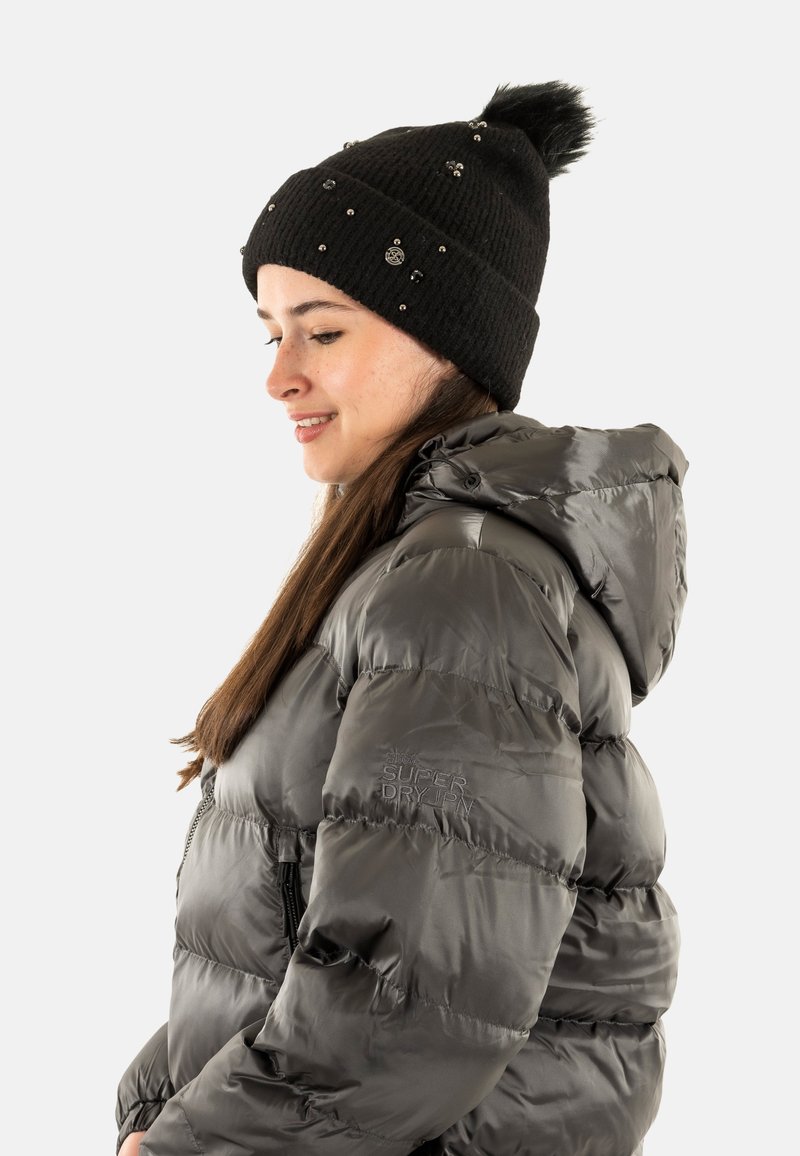 Cappello a maglia nera con pompon e decorazioni; giacca puffer grigia con chiusura a zip e cappuccio; logo del brand visibile sulla manica.