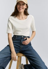 Witte geribbelde top met korte mouwen, gecombineerd met hoge taille wijde jeans van donkerdenim. Een lichtgekleurde pet en neutrale schoenen ronden de look af.