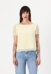 Blouse en chiffon jaune avec des manches bouffantes courtes et des accents floraux au niveau du col, assortie à un jean bleu. Texture lisse, coupe décontractée.
