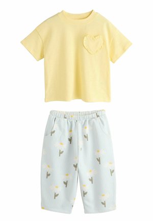 Hellgelbes Baumwoll-T-Shirt mit einem gerafften Herztasche, kombiniert mit blauen gestreiften Shorts mit weißen Gänseblümchen und gelben Akzenten.