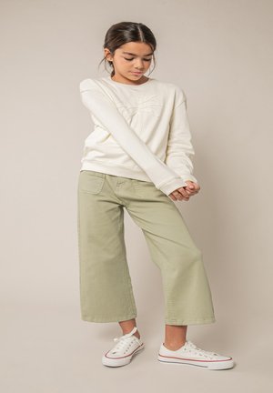 Fille aux cheveux foncés portant un sweat-shirt crème, un pantalon large vert clair et des baskets blanches, debout devant un fond beige.